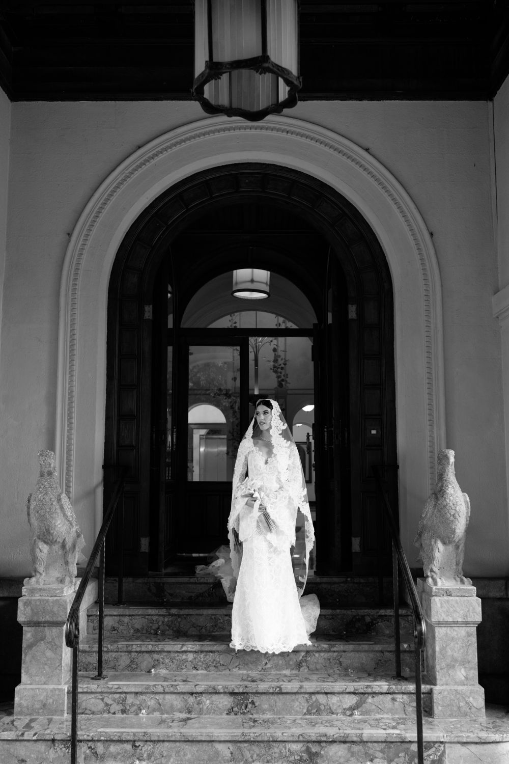 Black and white photo of a bride in white dress standing between classical stone pillars.