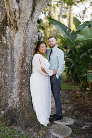 Wedding couple poses by large tree trunk in tropical garden setting.
