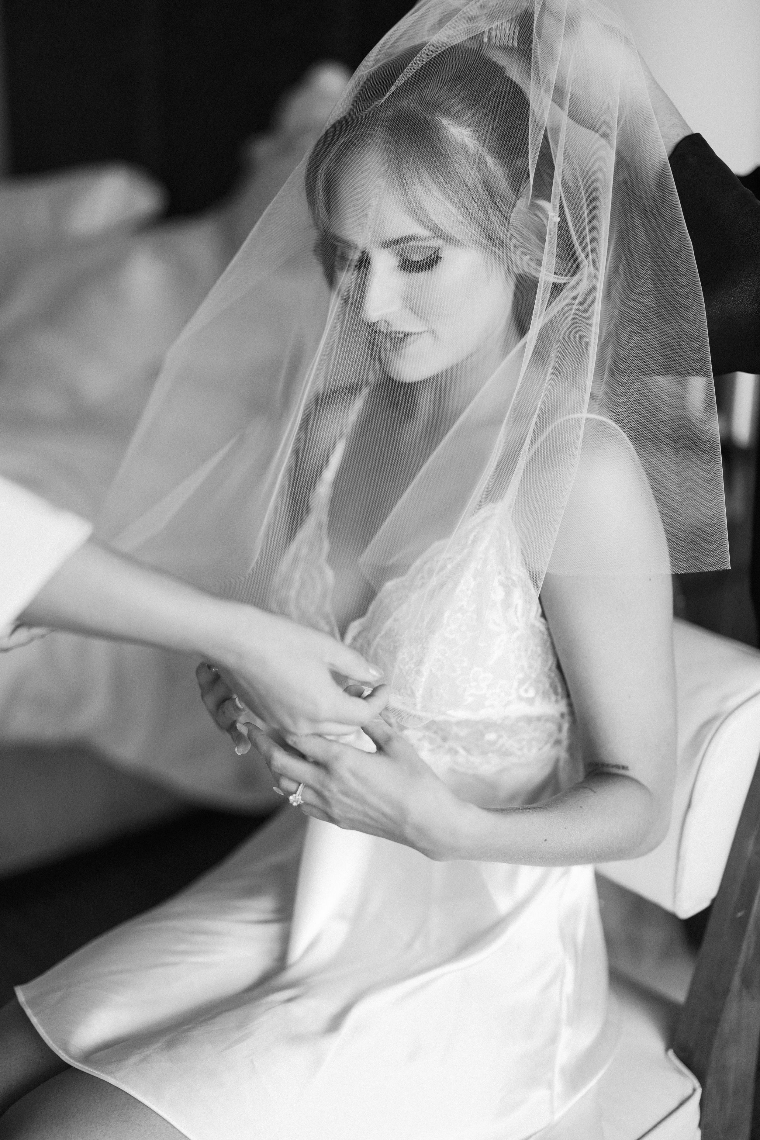 A bride in white wedding dress and veil looks down thoughtfully while sitting in a quiet moment of preparation.