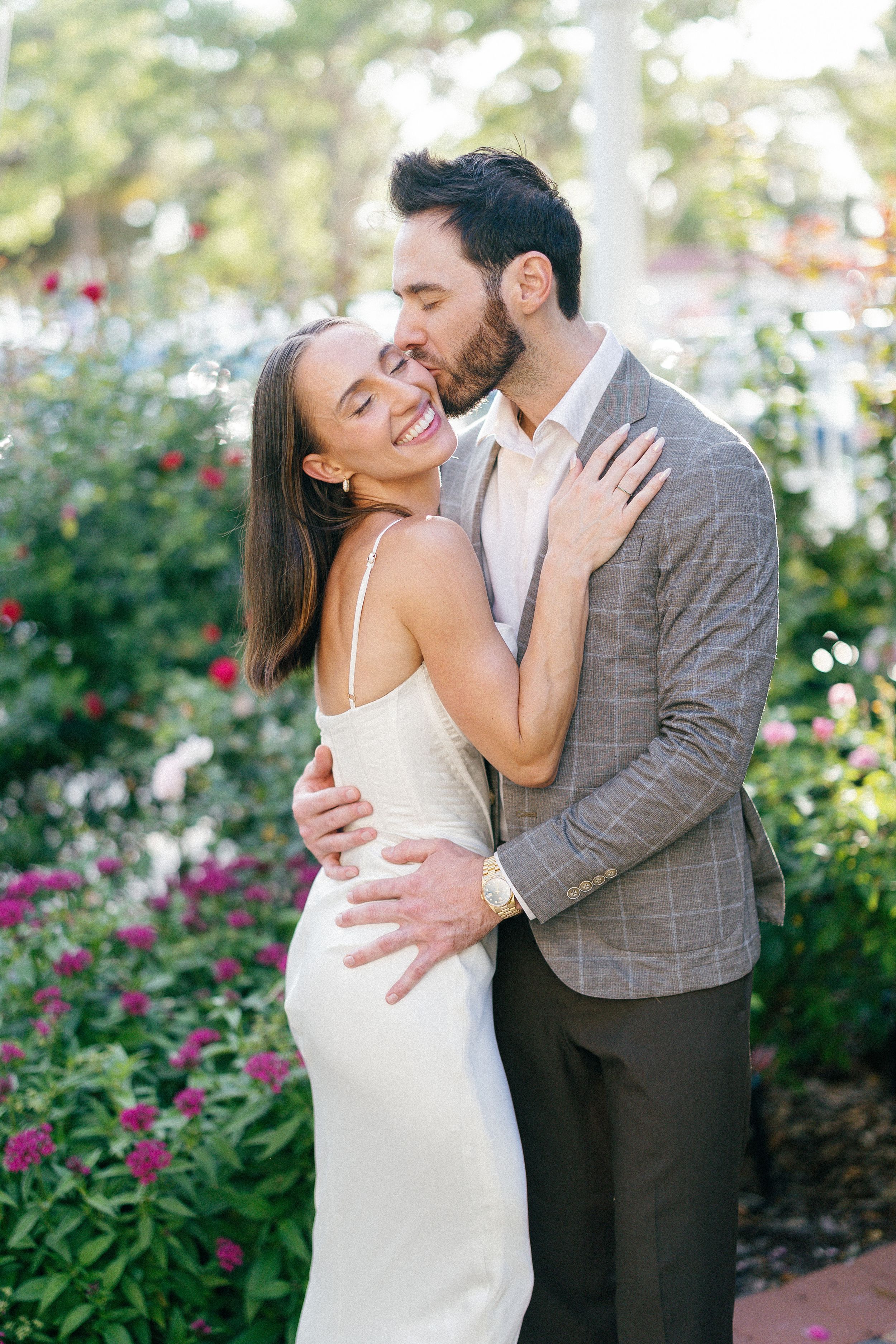 A romantic moment captured in a garden with blooming flowers, showing an embrace in elegant attire.