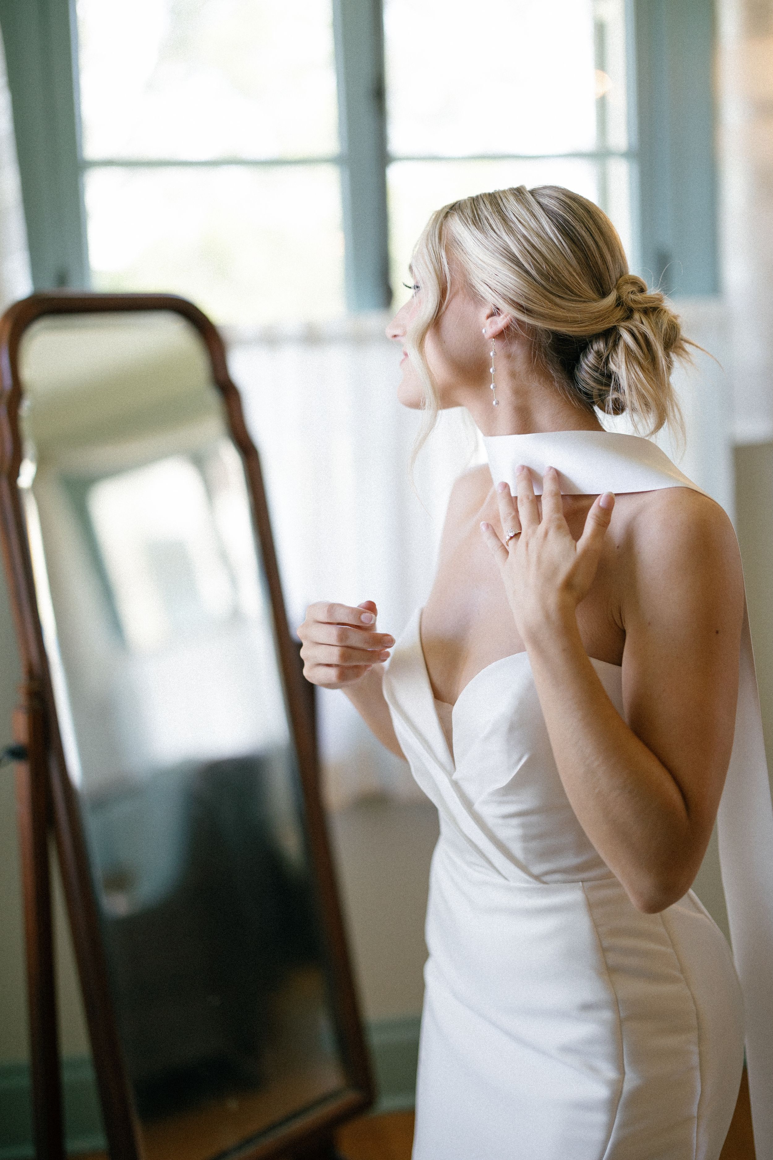 A woman in a white wedding dress stands before a full-length mirror in a sunlit room.