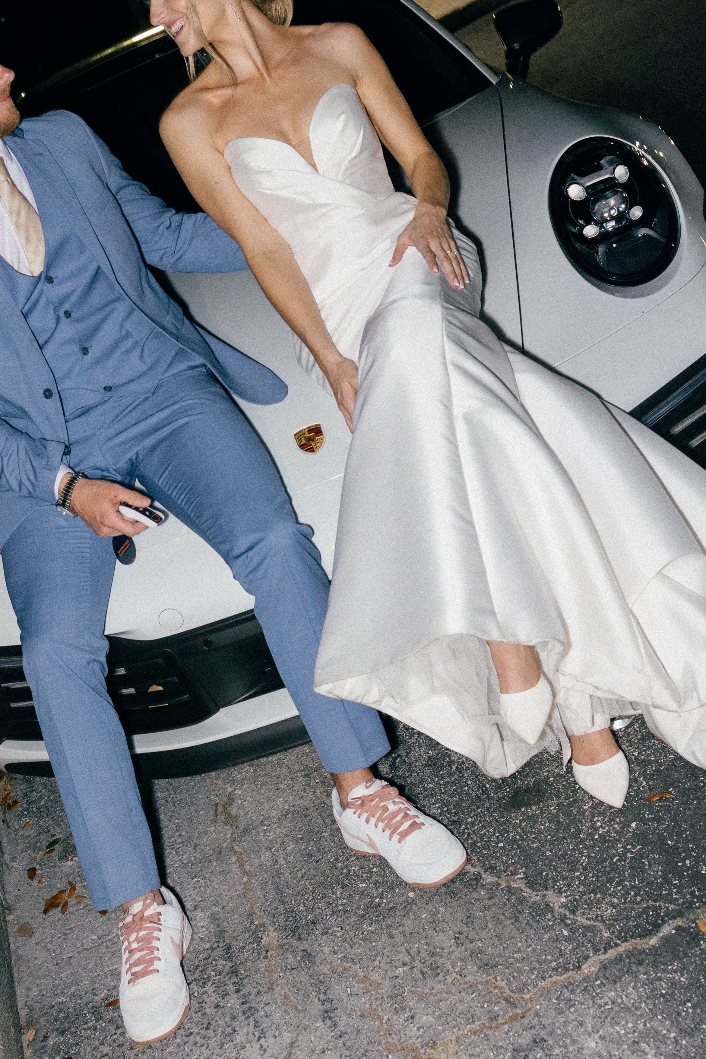 Modern couple in blue suit and white bridal gown showing off matching white sneakers next to a car.