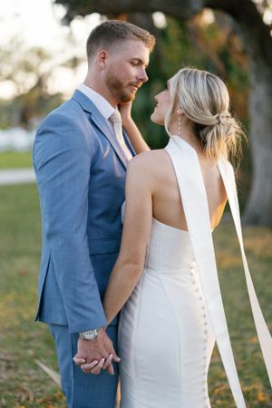 A bride in a sleek white dress and groom in a blue suit share an intimate moment outdoors during golden hour.