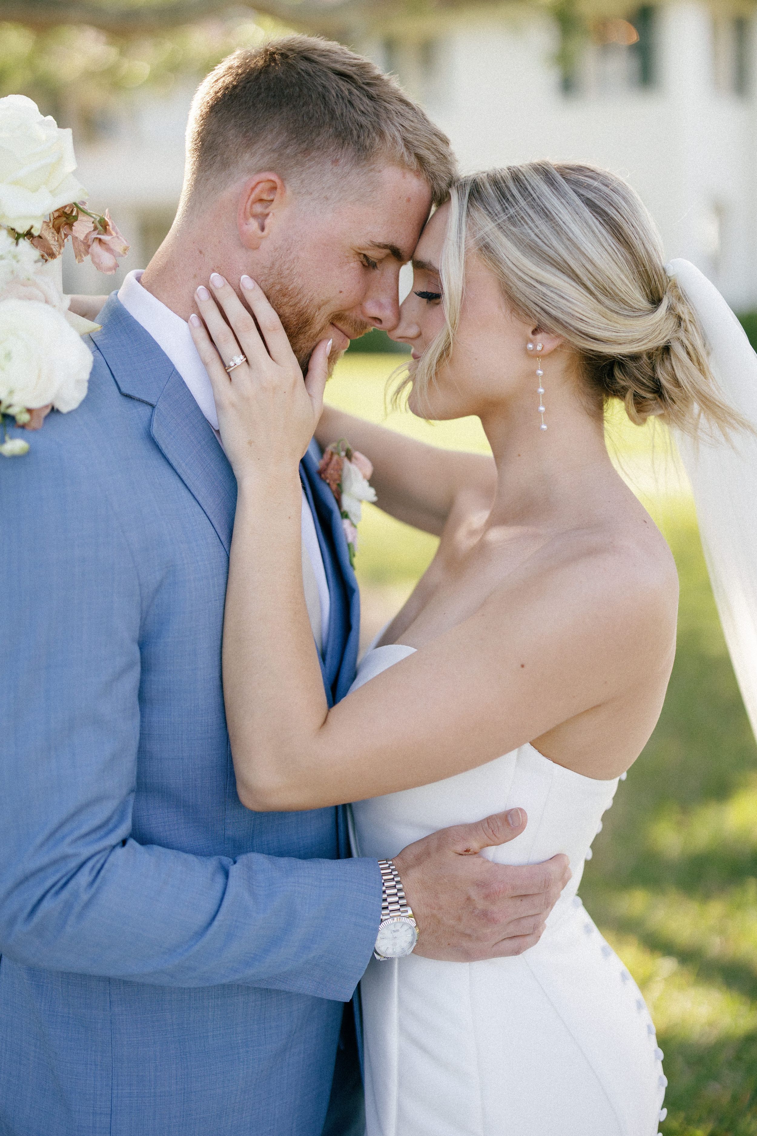 A tender moment between newlyweds in a light blue suit and white wedding dress outdoors in natural sunlight.