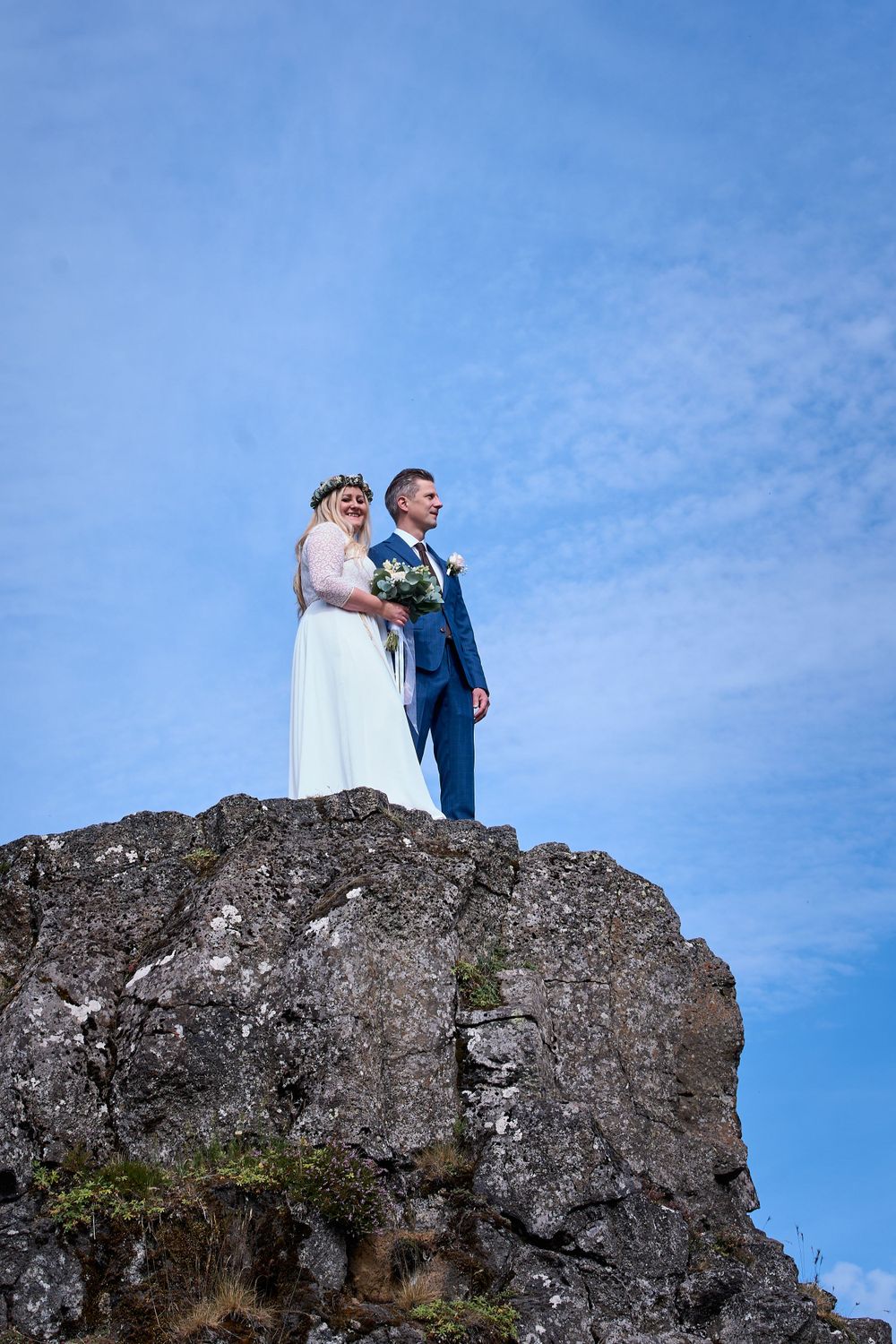 Newlyweds stand together on a dramatic rocky overlook against a bright blue sky.