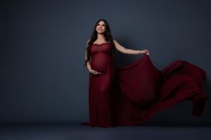 Dramatic maternity portraits in flowing burgundy dress against dark blue backdrop.