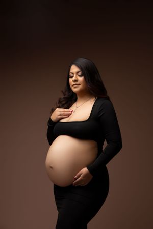 Elegant maternity portrait with expectant mother in black dress against dark brown backdrop.
