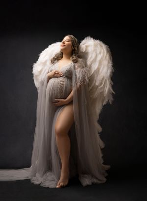 Artistic maternity photos with white angel wings and flowing sheer fabric against dark background.