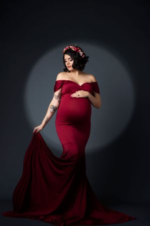 Dramatic maternity portrait featuring burgundy gown and floral crown against dark background.
