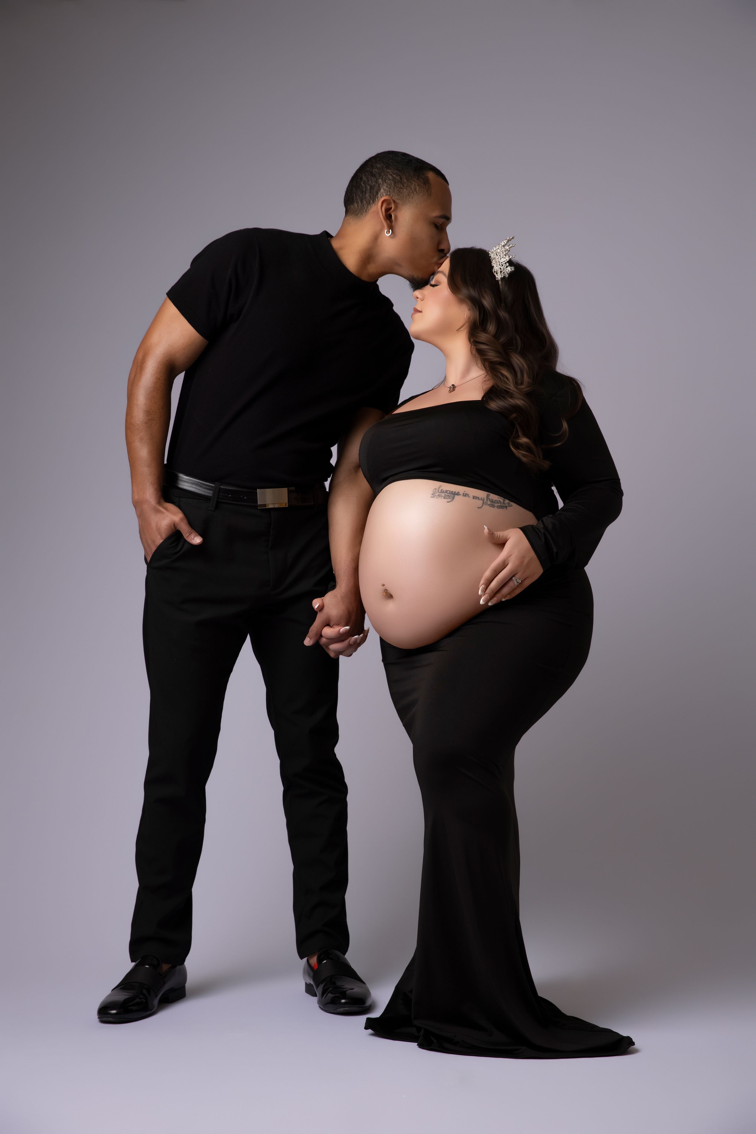 Intimate maternity photo of couple in black outfits sharing tender moment against grey backdrop.