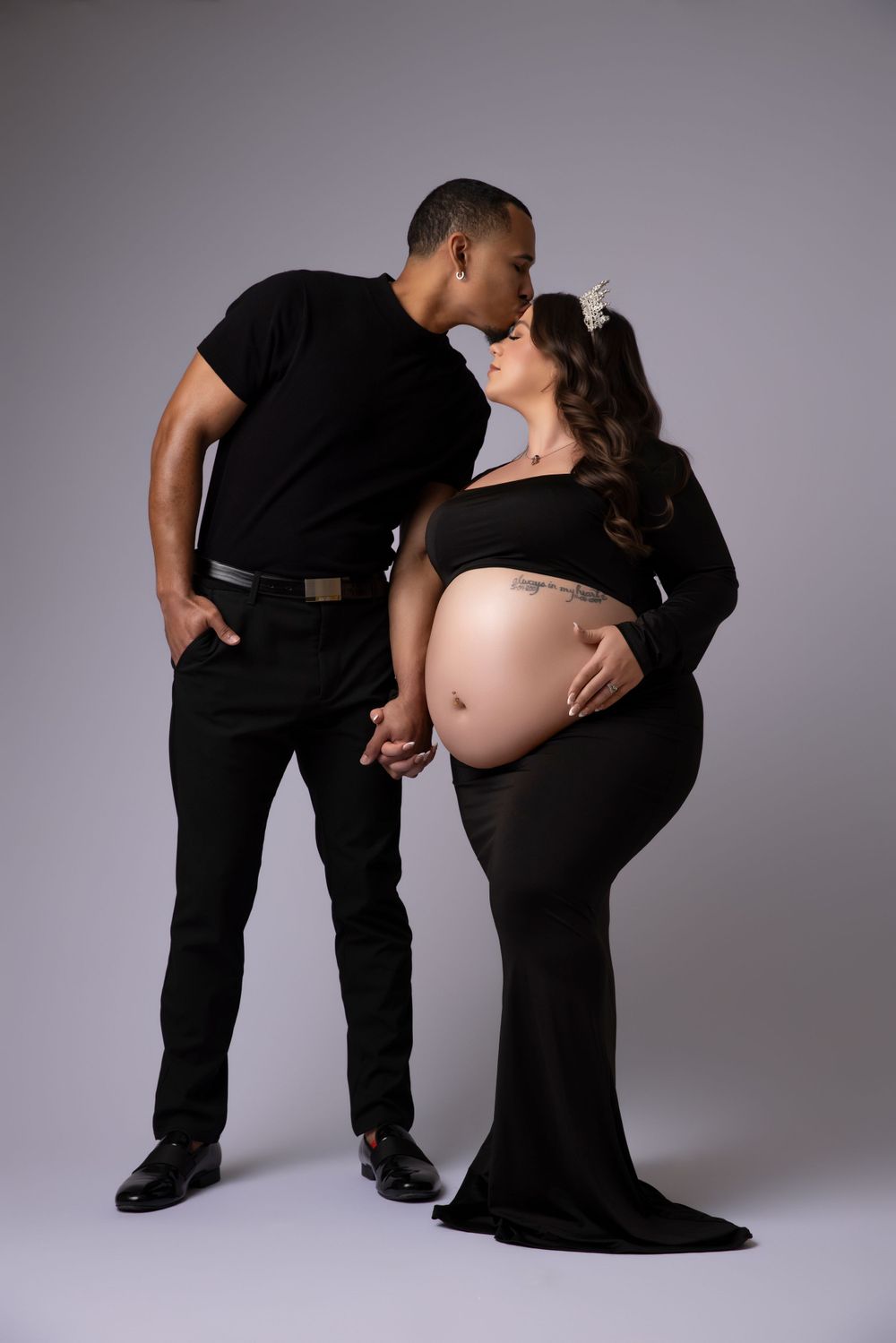 Intimate maternity photo of couple in black outfits sharing tender moment against grey backdrop.