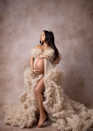 Maternity portrait featuring flowing cream dress and crown against textured brown background.