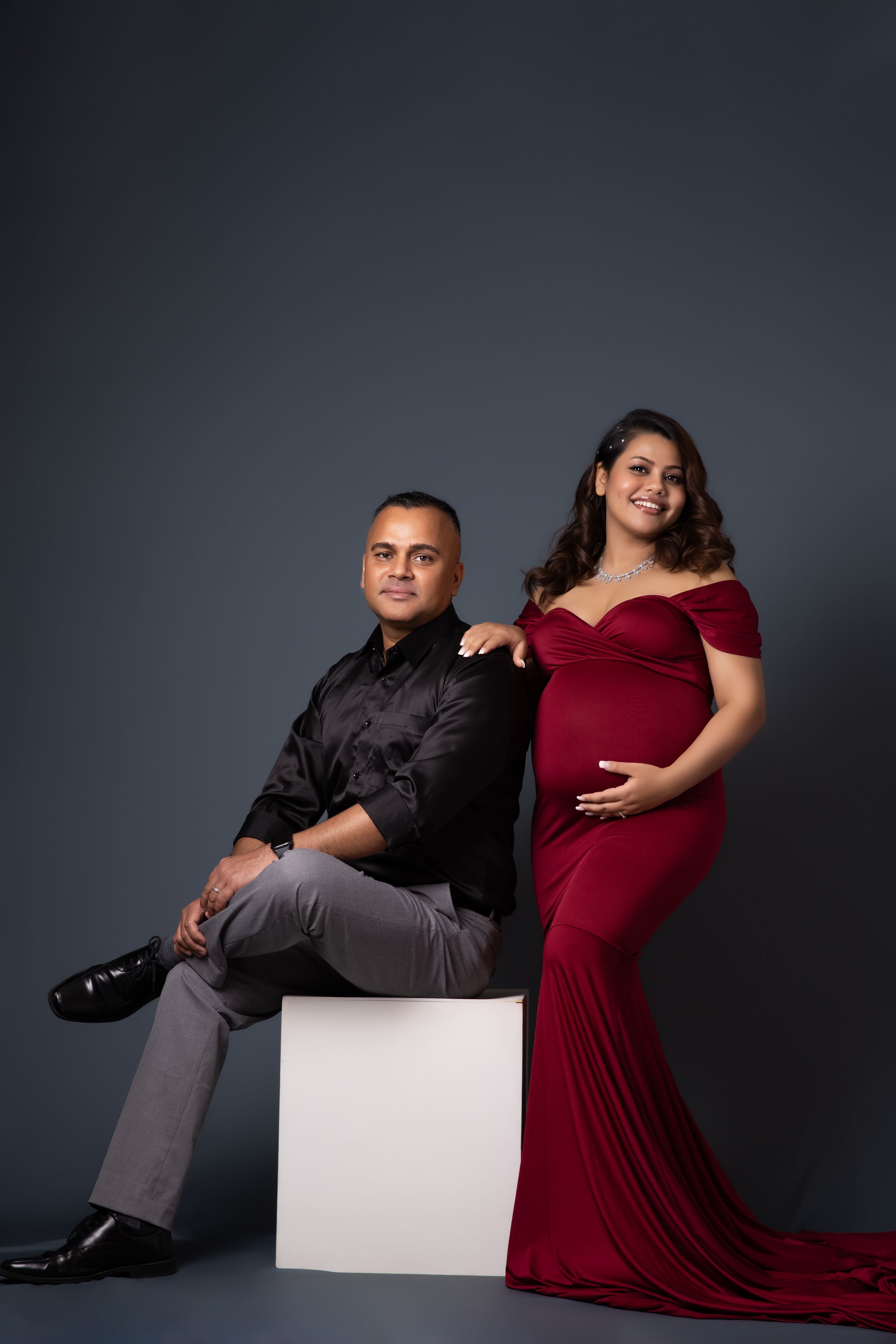 Couple's maternity session with red velvet dress and black attire against dramatic grey backdrop.