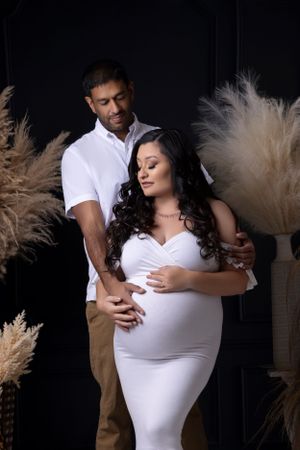 A romantic maternity photo featuring a couple embracing against a backdrop of pampas grass in a studio setting.
