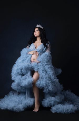 A maternity portrait with a flowing blue tulle gown and crystal crown against a dark background.