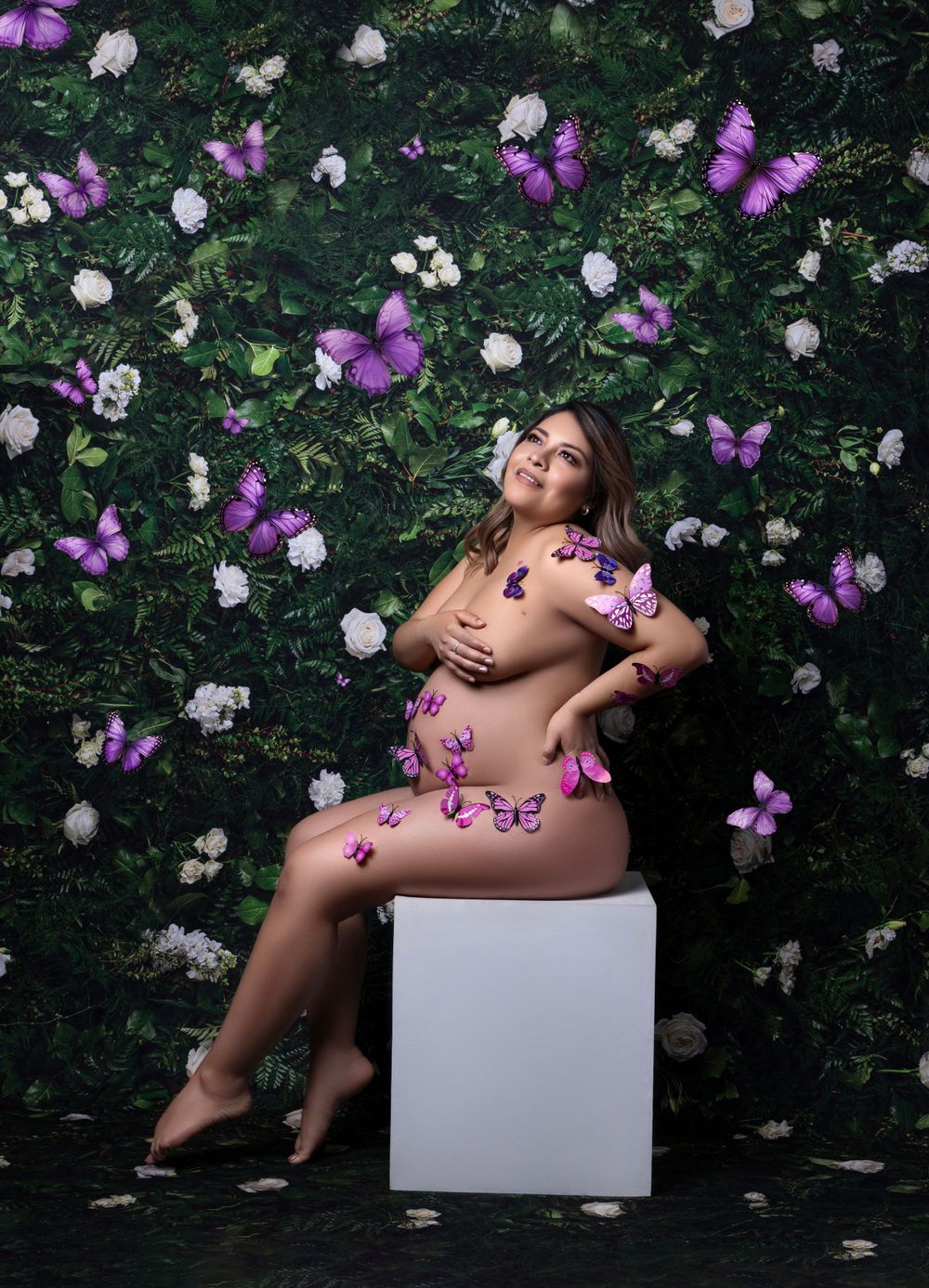 Artistic maternity photoshoot with purple butterflies and white flowers against a dark garden backdrop.