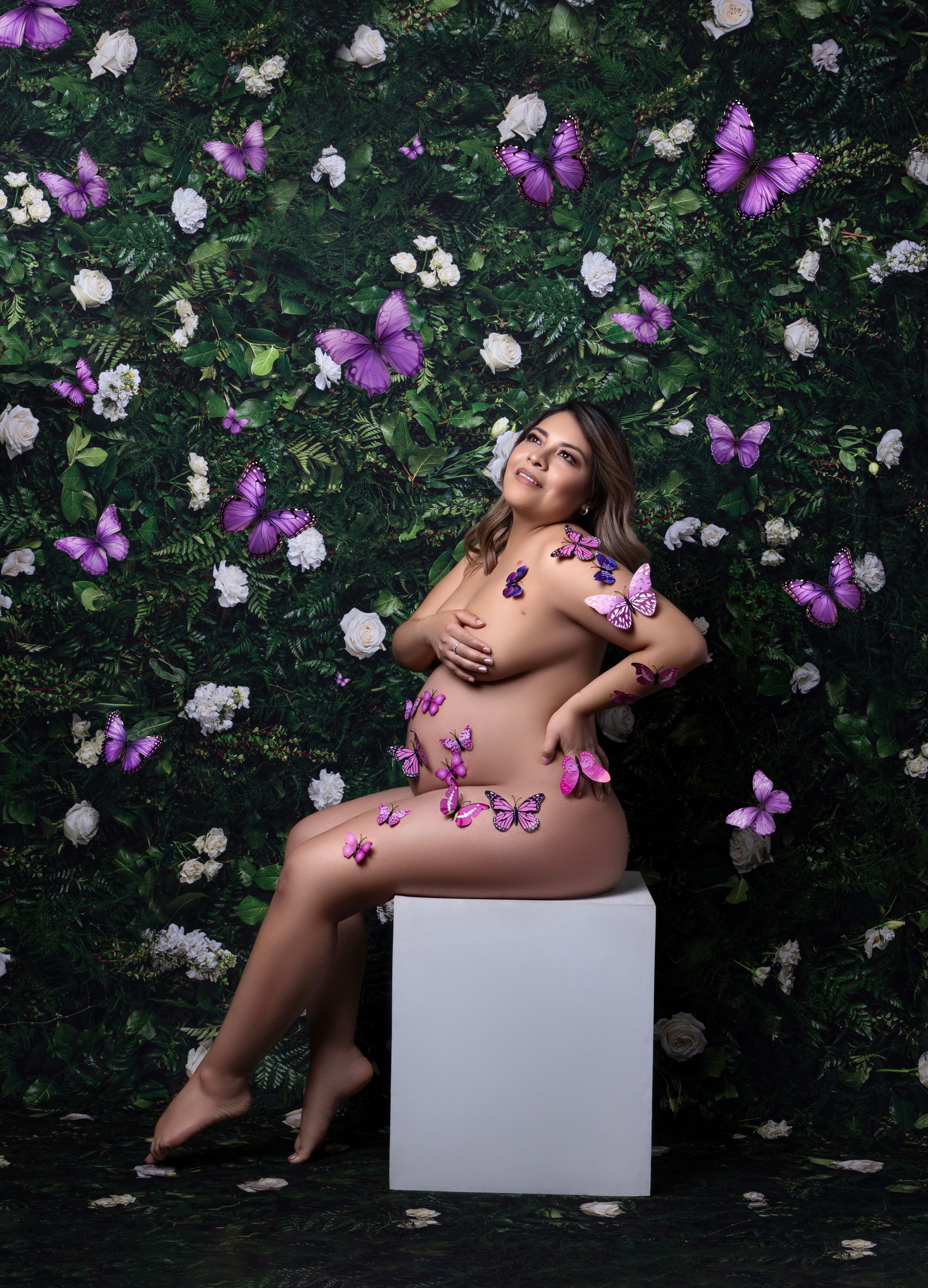 Artistic maternity photoshoot with purple butterflies and white flowers against a dark garden backdrop.