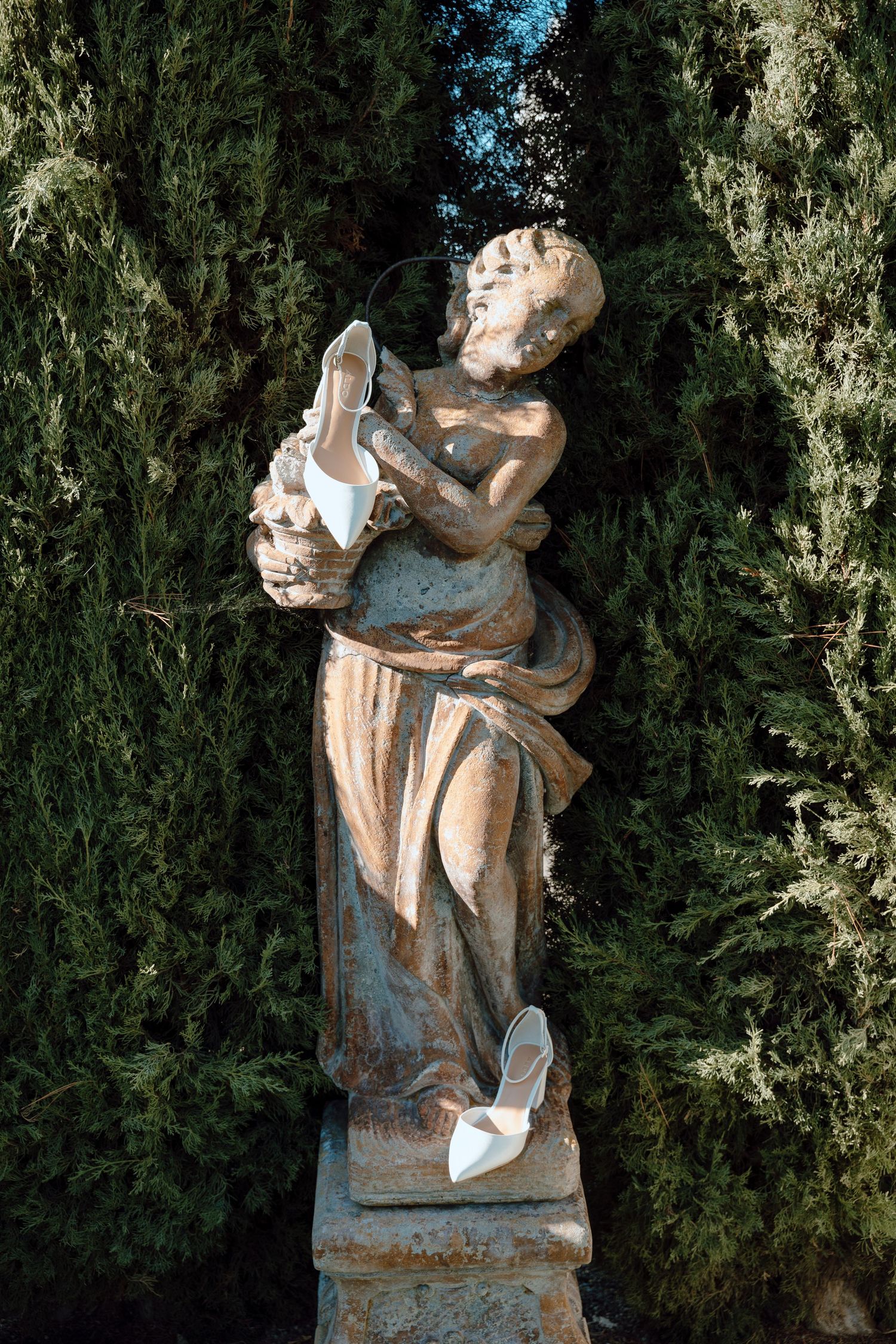 A classical stone statue adorned with wedding shoes stands among cypress trees in a garden setting.