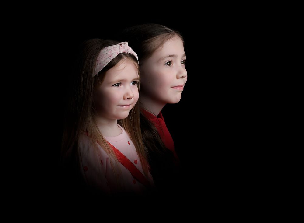 Sisters portrait side-lit dark moody studio image by Nicola in Woodside, capturing intimate connection with dramatic lighting, timeless.