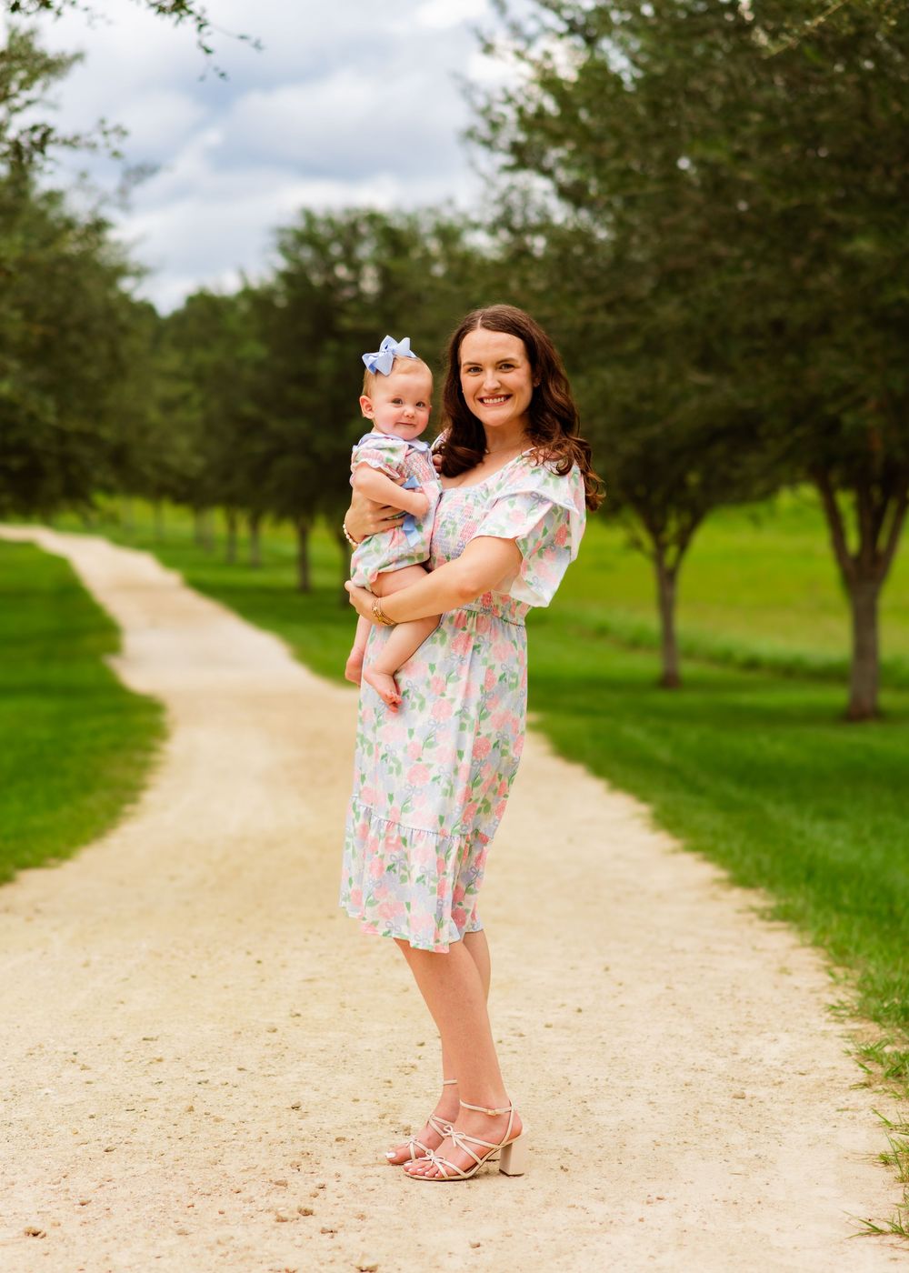 A smiling woman in a floral dress holds a baby girl with a blue bow on a scenic tree-lined gravel path.