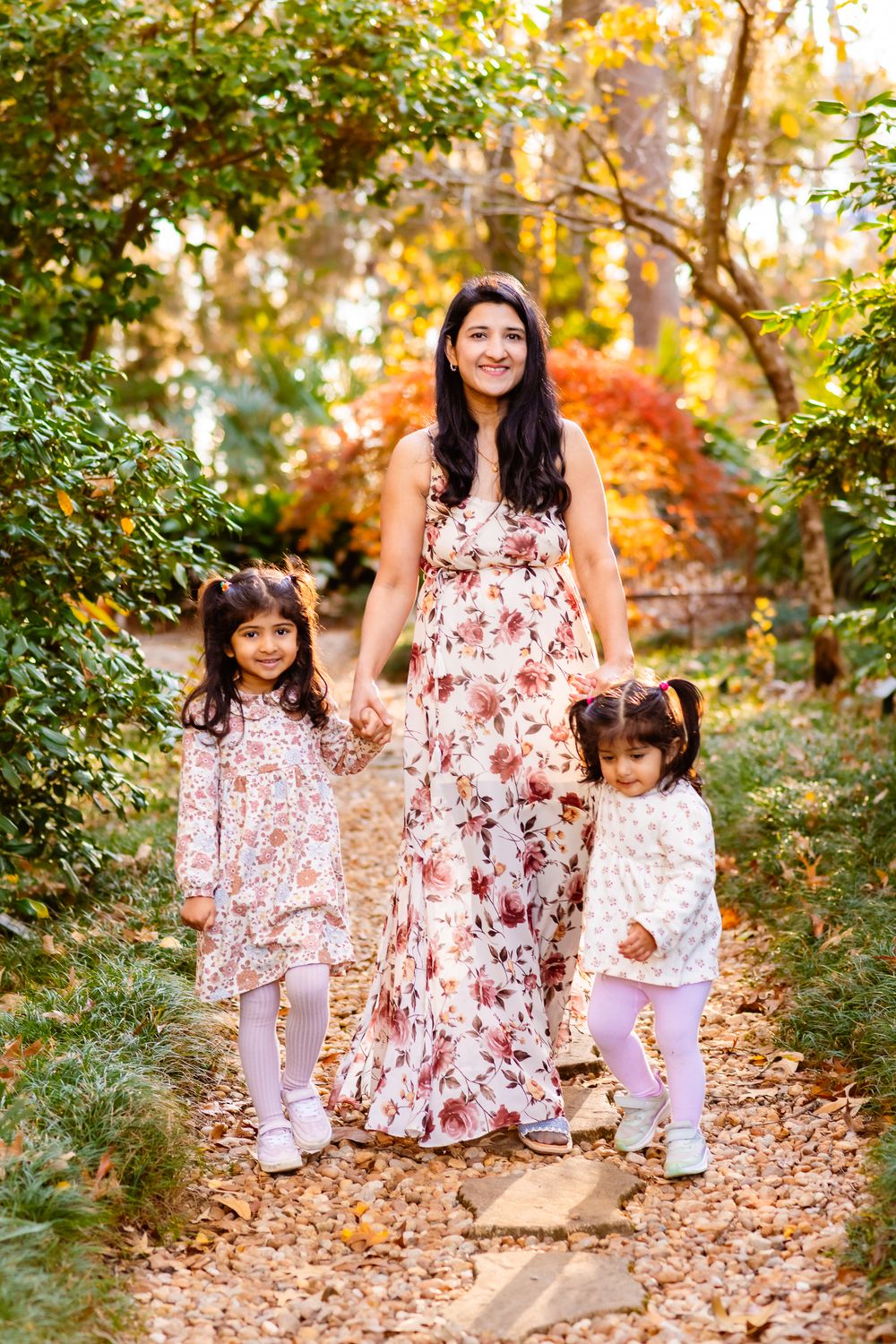 A family in matching floral dresses walks through a sunlit autumn forest path covered in fallen leaves.