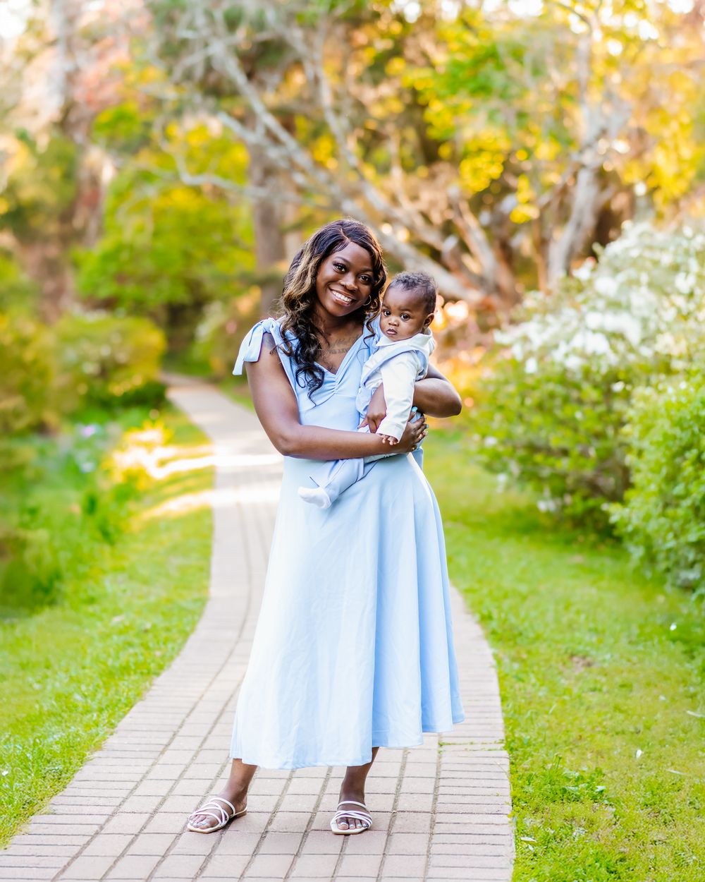 A smiling woman in a light blue dress holds her baby on a scenic garden path surrounded by lush greenery.