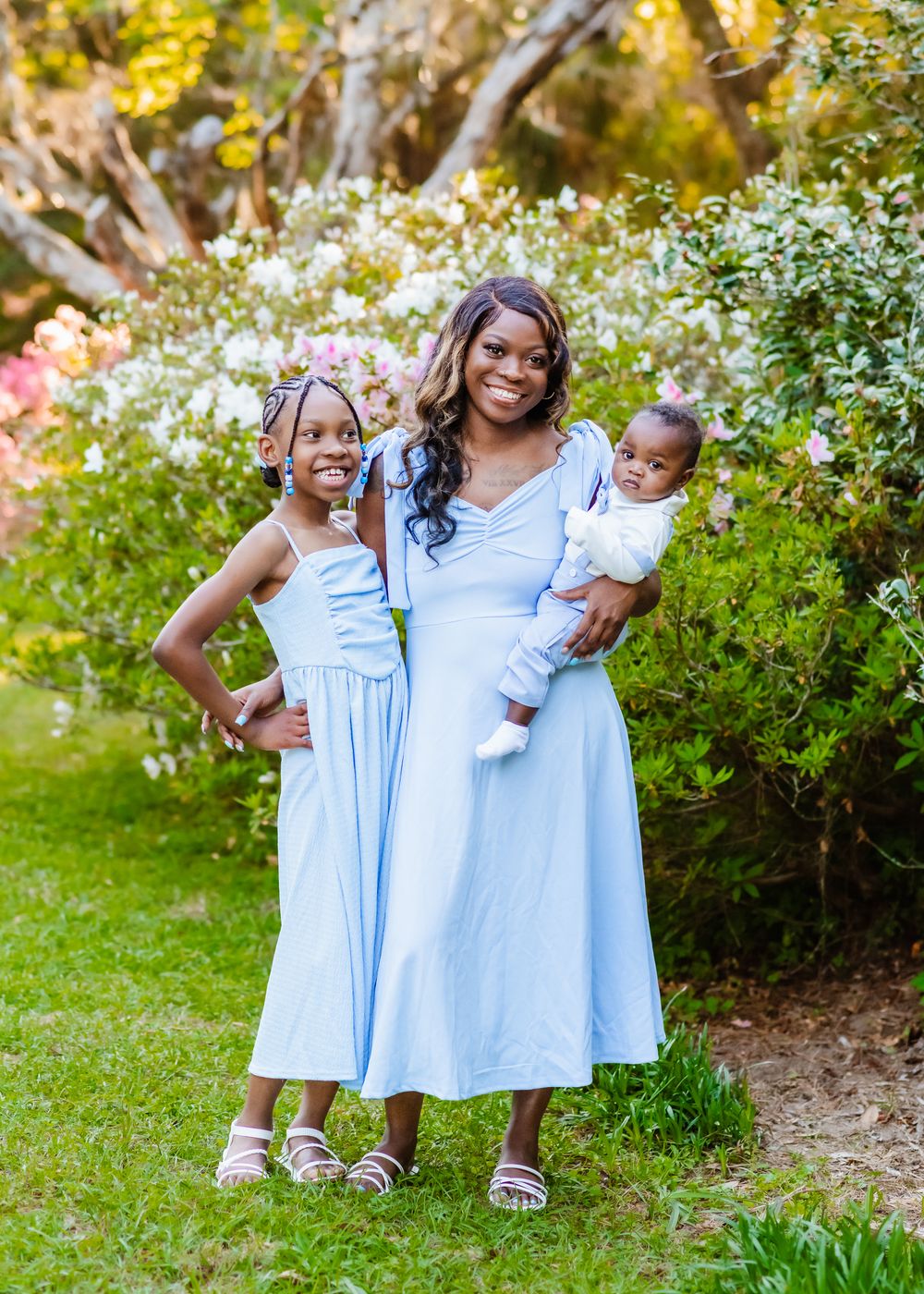 A mother in a light blue dress holds her baby while posing with her daughter in a garden.