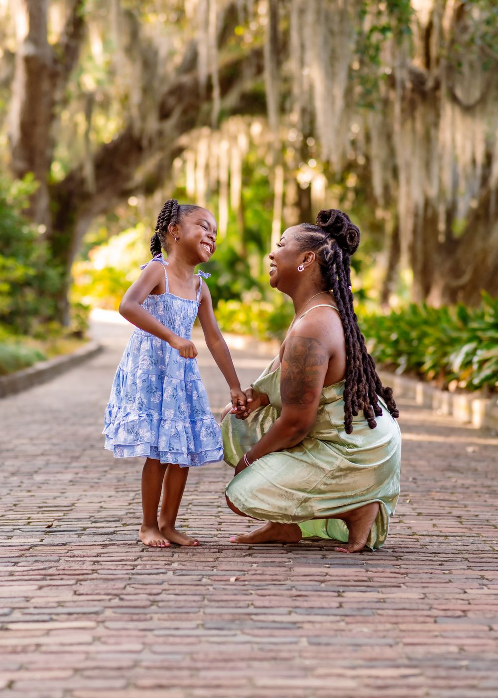 A mother kneels on a brick path holding hands with her young daughter in a lush green park setting.