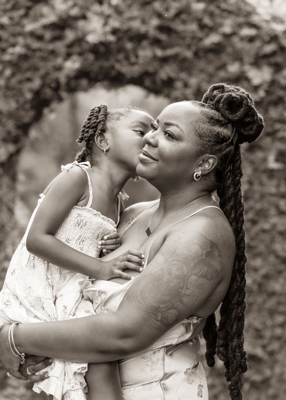 Black and white photo of a woman holding a young girl who kisses her cheek, outdoors near ivy-covered wall.
