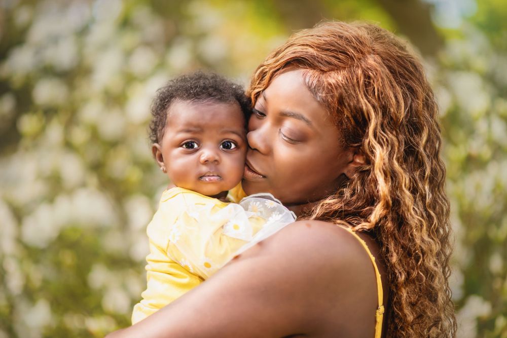 Parent with long curly hair embraces baby wearing yellow outfit outdoors with soft bokeh lighting.