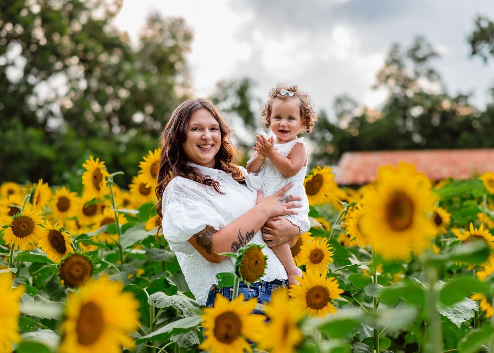 A mother holds her toddler in a vibrant sunflower field, both smiling joyfully on a cloudy day.