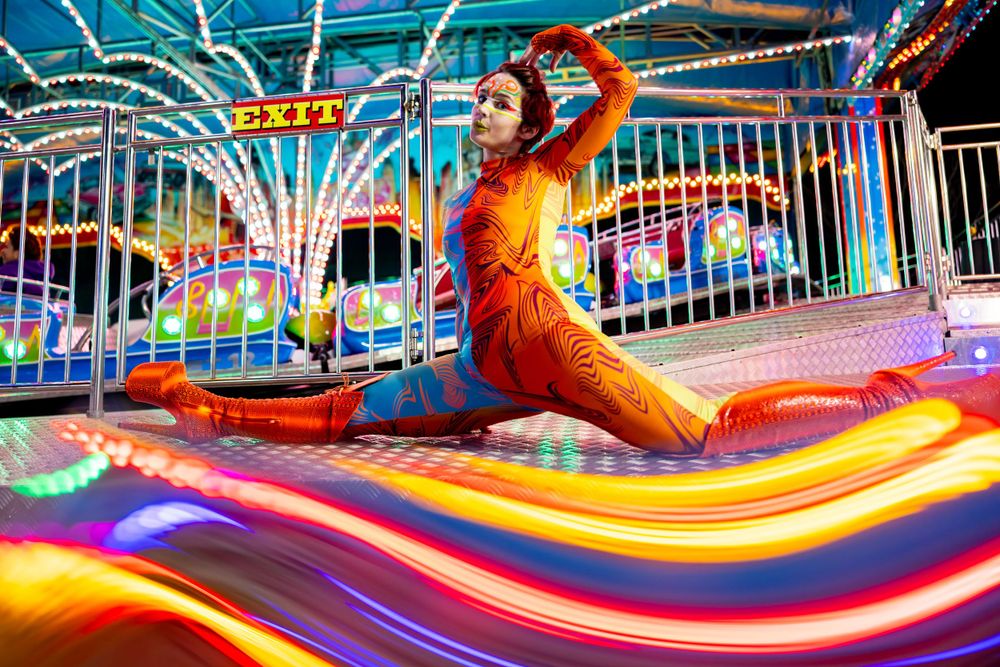 Performer in vibrant orange costume strikes artistic pose on colorful carnival ride.