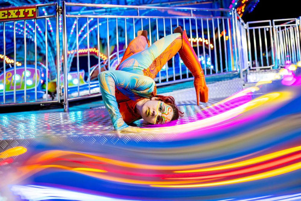 Performer in colorful outfit does acrobatic moves on carnival ride with neon light trails.