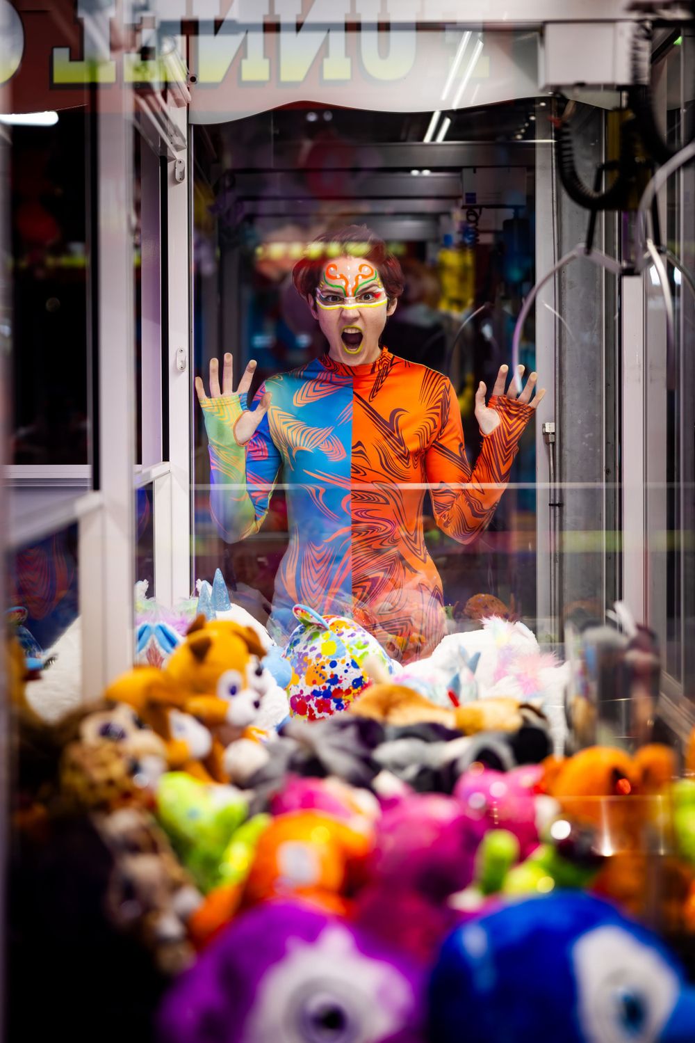 Energetic performer poses expressively in claw machine filled with colorful plush toys.