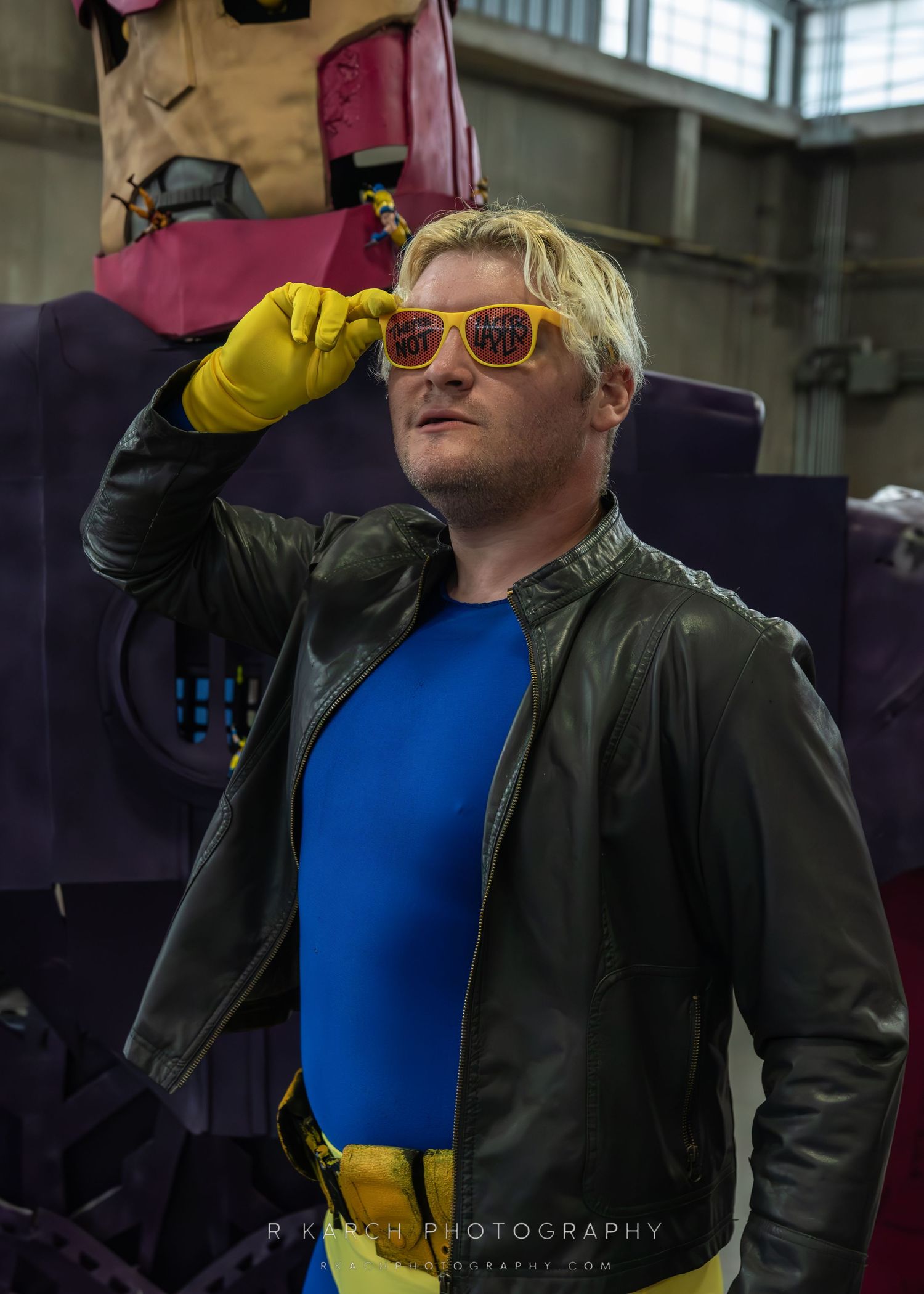 A performer in a leather jacket and sunglasses strikes a dramatic pose against an industrial background.