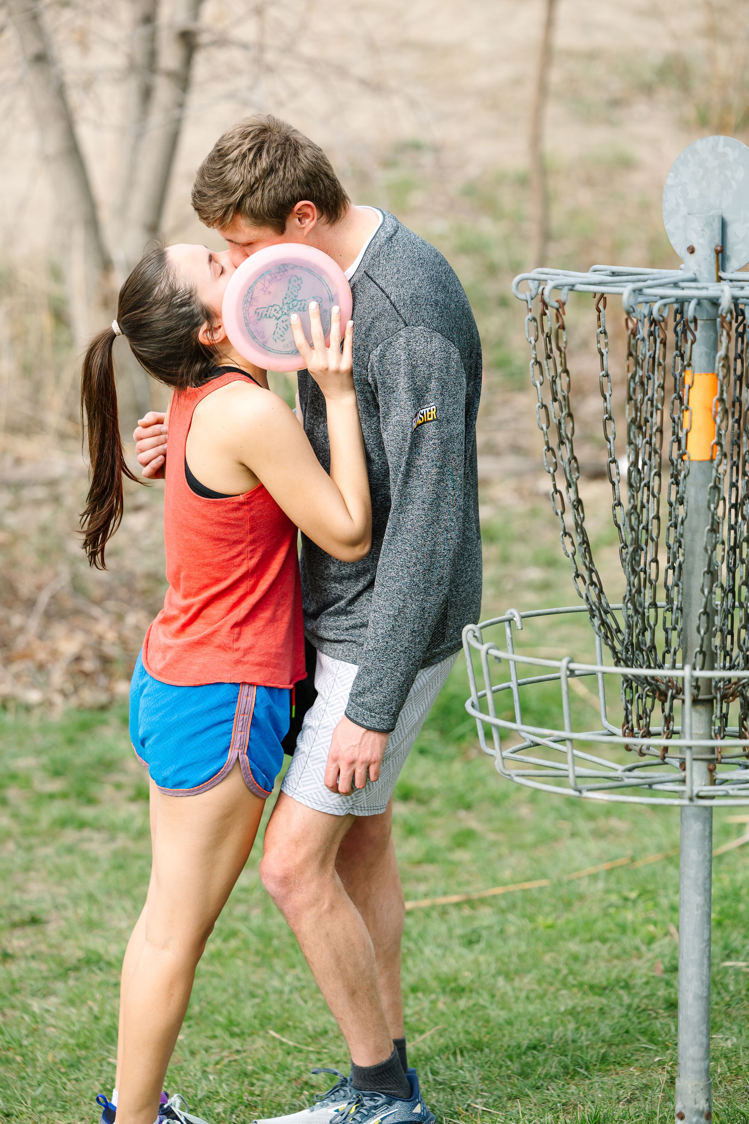 A couple shares a romantic kiss while playing disc golf on a course with a basket visible in the background.