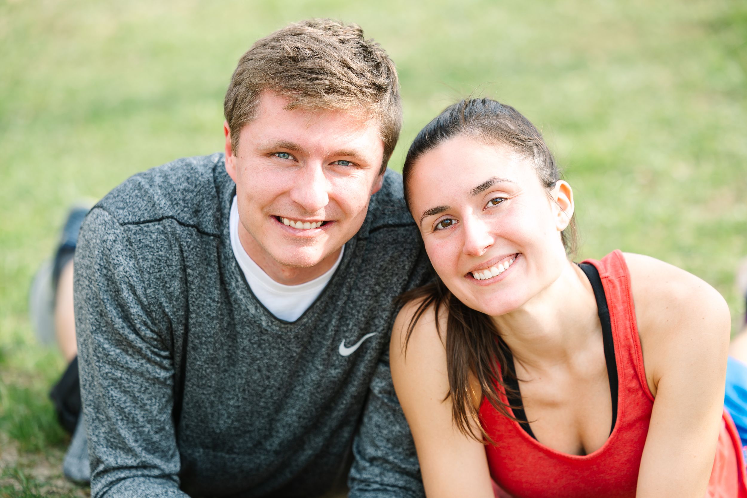 A couple wearing athletic wear sits together outdoors on grass on a sunny day.