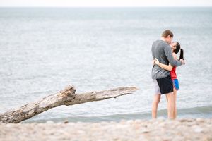 Two people share a romantic moment on a beach near driftwood and waves during an engagement photoshoot.
