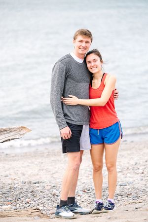 A couple in athletic wear embraces while standing on a beach shore on a cloudy day.