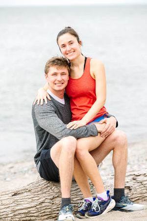 A couple in casual athletic wear sits together on a log by the water on a bright overcast day.