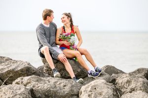 A couple sits together on coastal rocks, smiling at each other while wearing casual summer clothing.