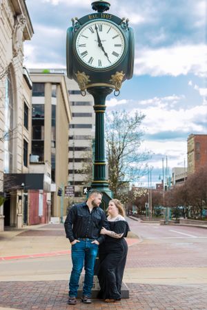 Couple poses romantically under vintage street clock in urban setting.