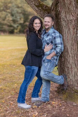 A couple poses together against a large tree trunk in a park setting.