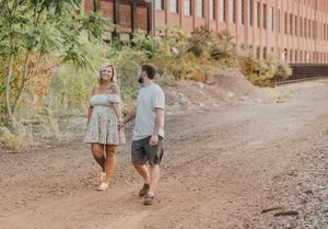 Couple walking and laughing together on a dirt path near brick buildings.