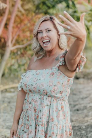 Excited person showing off engagement ring in floral dress against nature background.