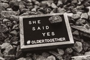 Black letter board on rocks displaying 'She Said Yes #OlderTogether'.