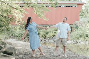 Couple holding hands and laughing by covered bridge in coordinated outfits.