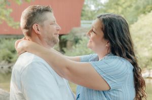 A romantic couple embraces outdoors near a red barn on a sunny day.