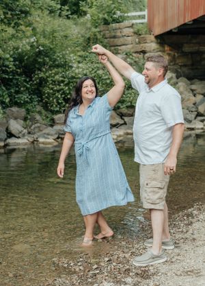 A couple dancing playfully in shallow creek water near a covered bridge.