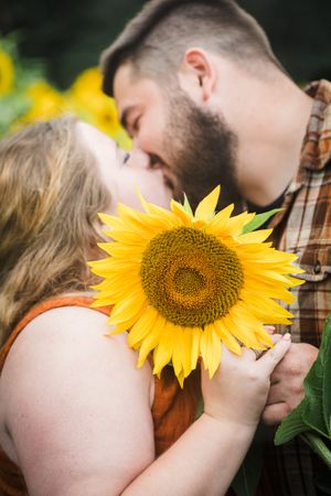 A romantic kiss behind a bright yellow sunflower in a garden setting.